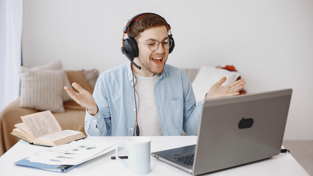 Muškarac drži online čas preko laptopa uz slušalice, knjigu i beleške na stolu